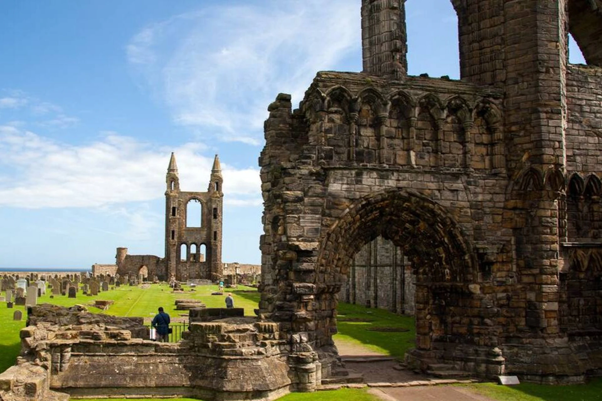 Ruins of an ancient stone abbey with tall arches and gravestones on green grass under a blue sky with clouds.