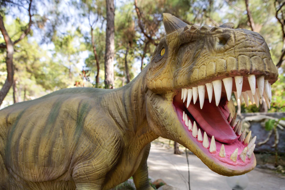 Close-up of a dinosaur statue with an open mouth displaying sharp teeth, set against a backdrop of trees and a walkway.