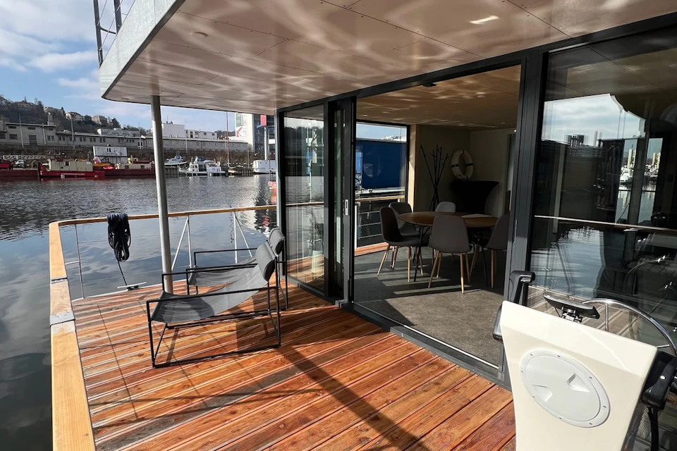 A modern houseboat with a wooden deck, outdoor chairs, and glass doors leading to an interior dining area by a harbor.
