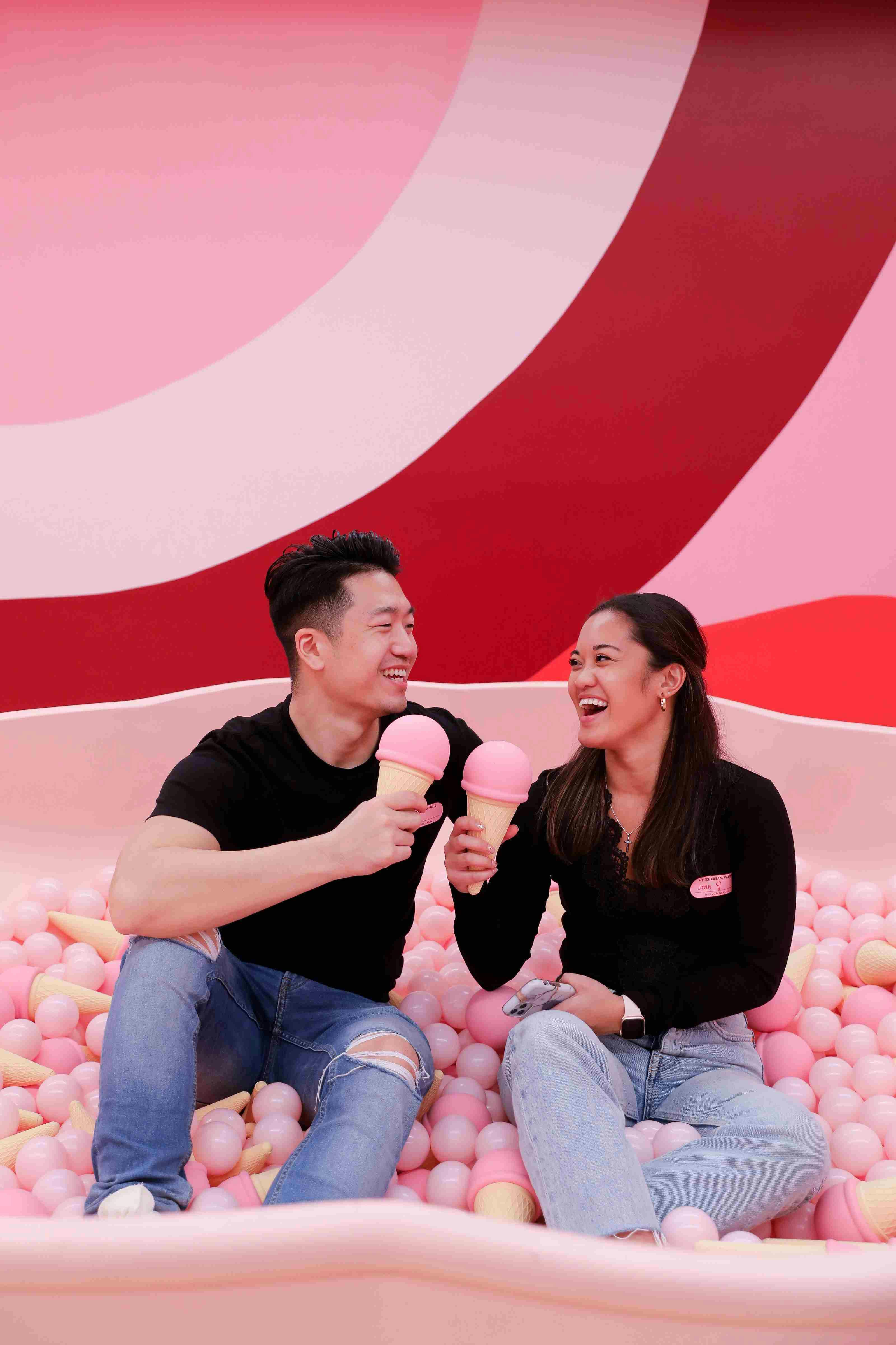 Two people sitting in a ball pit, smiling and holding ice cream cones against a colorful red and pink background.