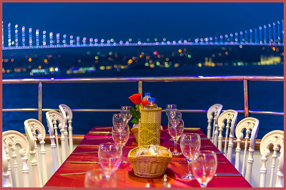 A table set for dinner with wine glasses and a basket under a night sky, with a lit-up bridge in the background.