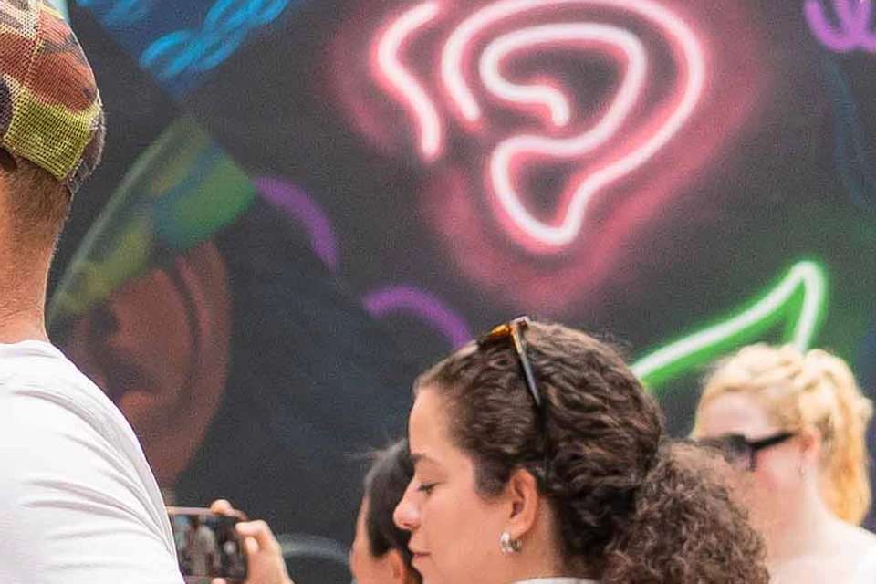 People observe a vibrant, graffiti-covered wall in an outdoor urban space. Some take photos while others engage in conversation.