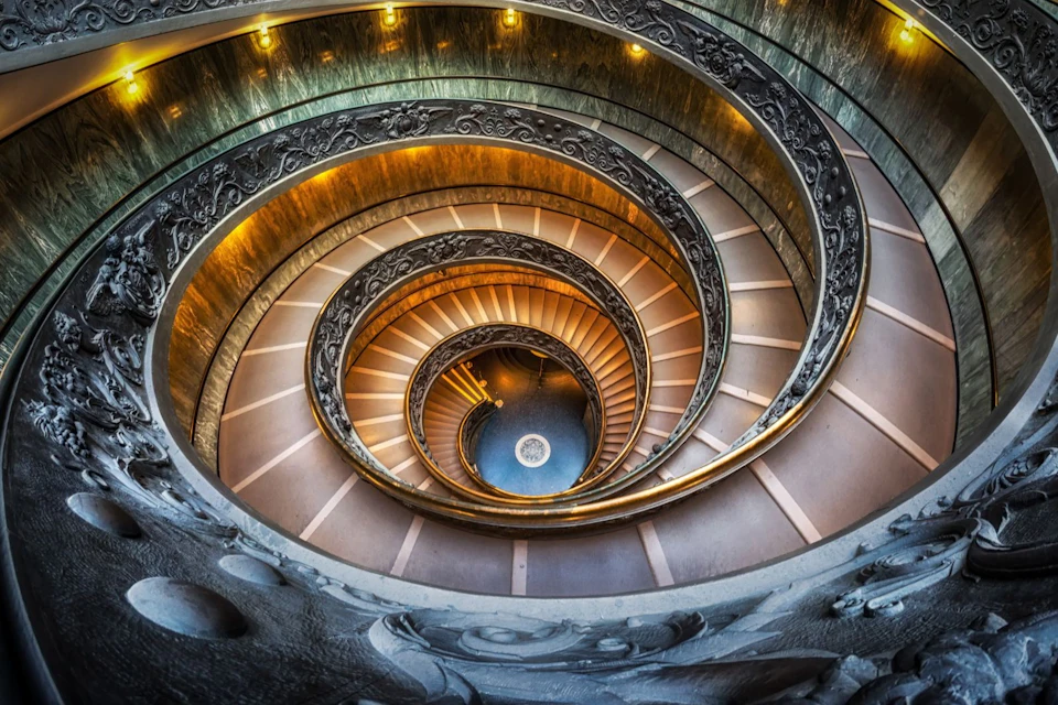 Spiral staircase with ornate railings and warm lighting, viewed from above, leading down to a blue floor with a circular pattern.