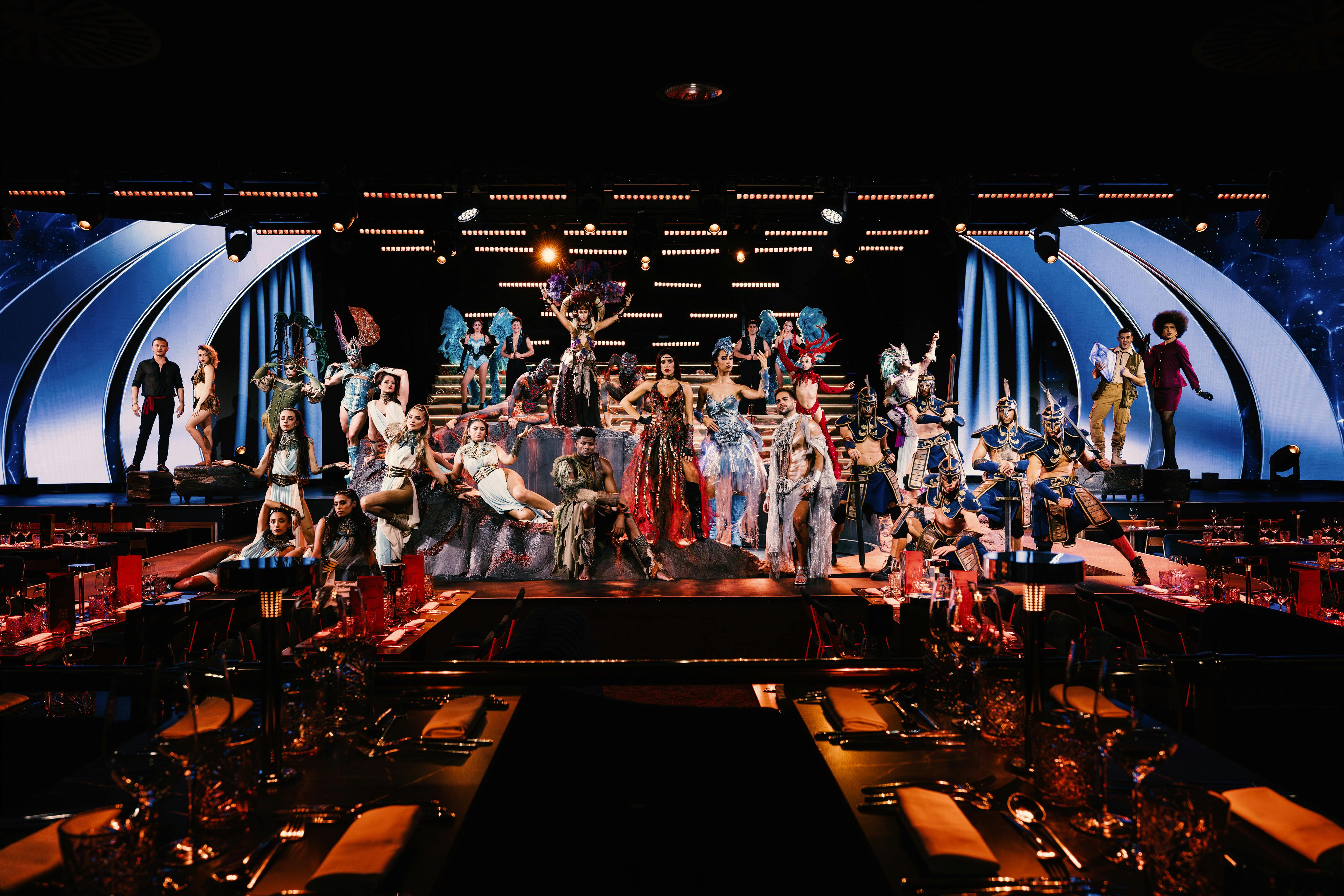 Танцоры в сложных костюмах выступают на сцене с драматическим освещением, смотря из обеденной зоны с накрытыми столами.