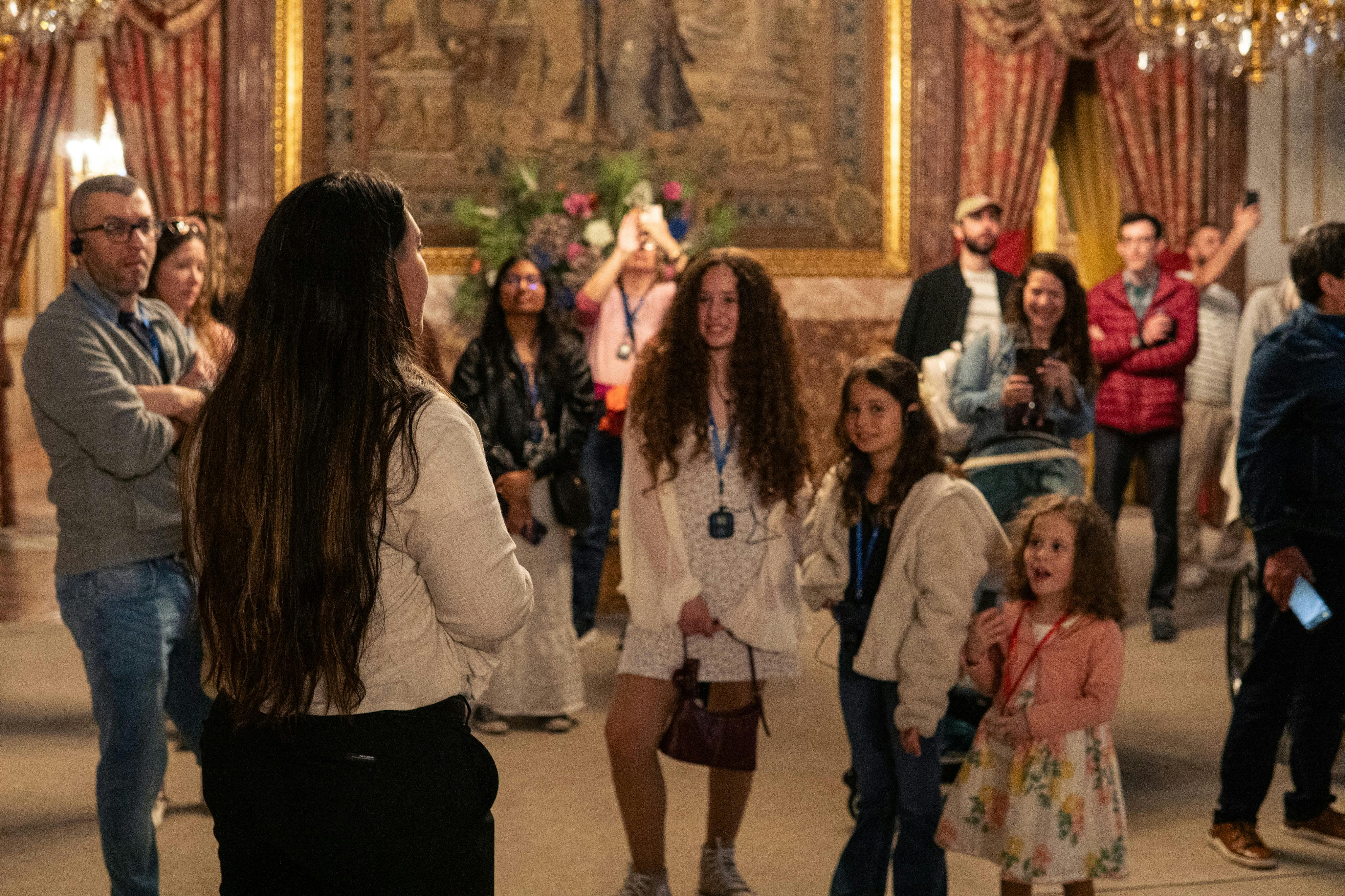 A group of people, including children, standing in an ornate room with tapestries and chandeliers visible in the background.