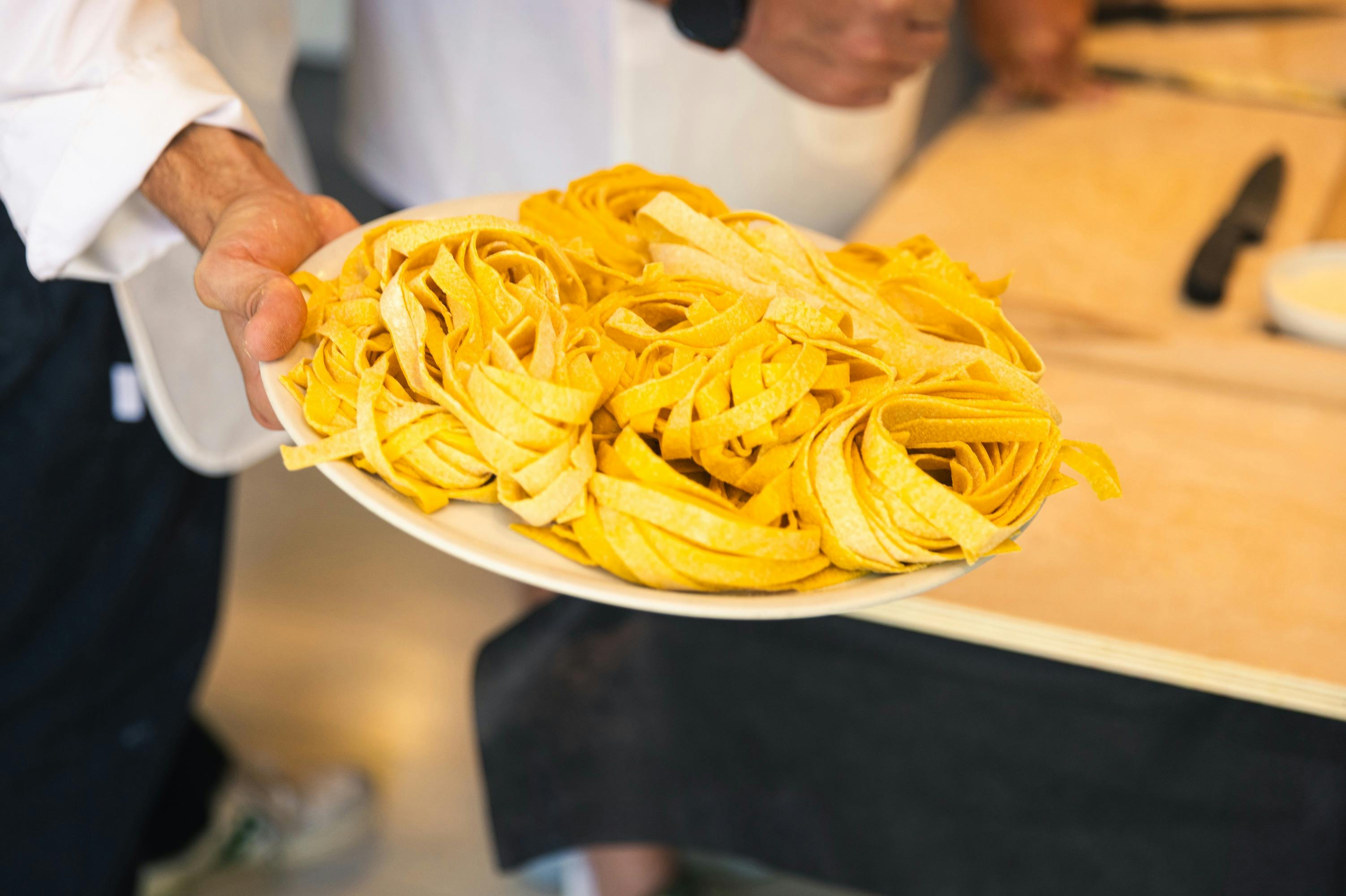 A plate of fresh yellow pasta nests held by a person with a table and another person visible in the background.
