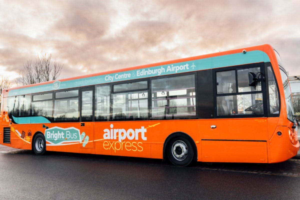 En ljusorange och blågrön buss med "Bright Bus airport express" på sidan, parkerad på en väg med molnig himmelbakgrund.