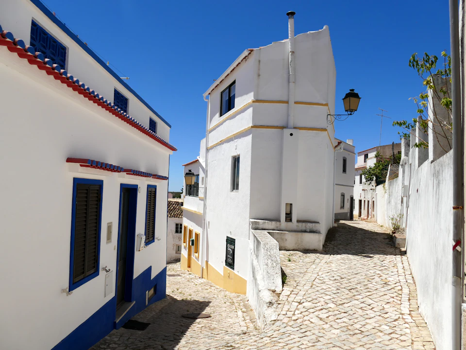 Narrow cobblestone street with white buildings featuring blue and yellow accents, under a clear blue sky.