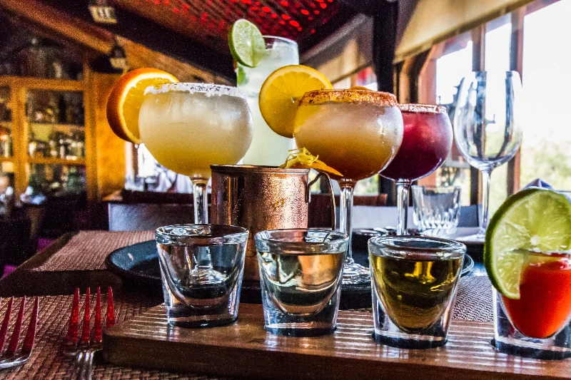 A variety of colorful cocktails and shots on a wooden board inside a dimly lit bar or restaurant.