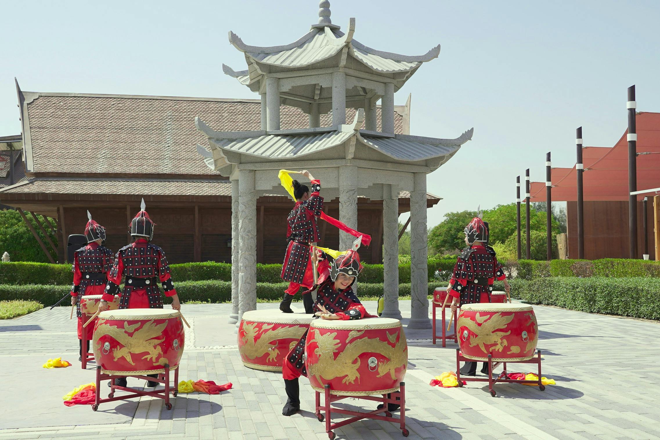 Τέσσερις άνθρωποι με παραδοσιακή ενδυμασία παίζουν διακοσμημένα τύμπανα σε εξωτερικούς χώρους κοντά σε ένα πέτρινο περίπτερο με δέντρα στο παρασκήνιο.