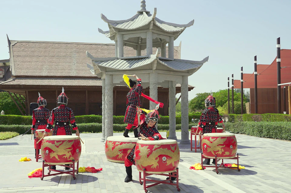 Four people in traditional attire playing decorated drums outdoors near a stone pavilion with trees in the background.