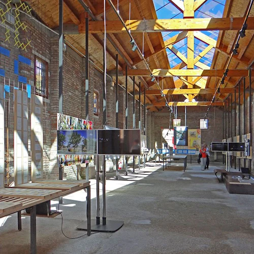 Rymlig utställningshall med träbjälkar, takfönster och olika displayer på bord och väggar. Två personer tittar på utställningar.