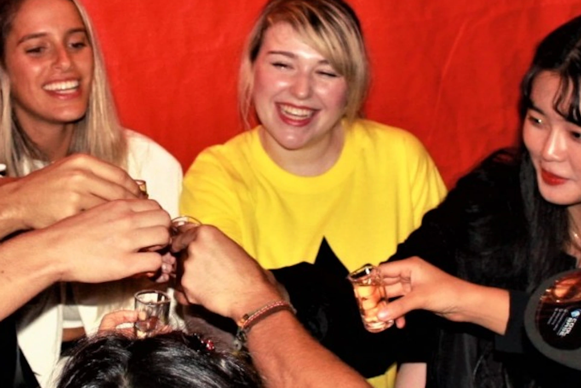 A group of people sitting around a table, smiling and raising their glasses in a toast, against a red background.