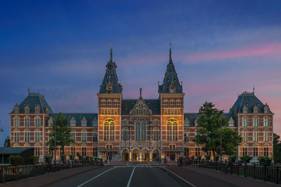 A historic building with large windows and towers at dusk, illuminated by lighting with a clear sky in the background.
