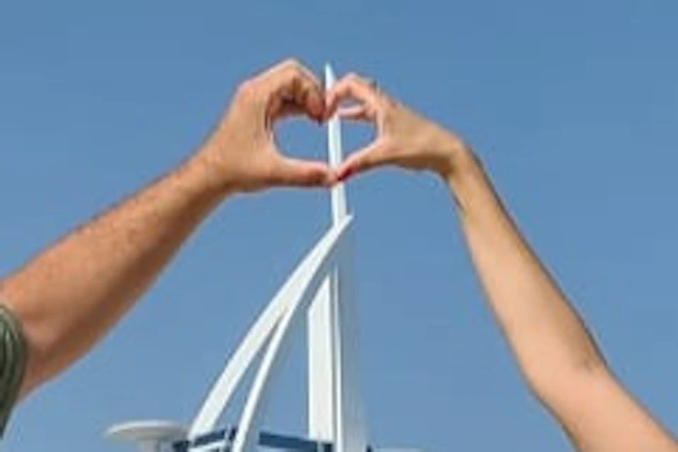 Two people stand on a sandy beach forming a heart shape with their hands. Modern buildings are visible in the background.