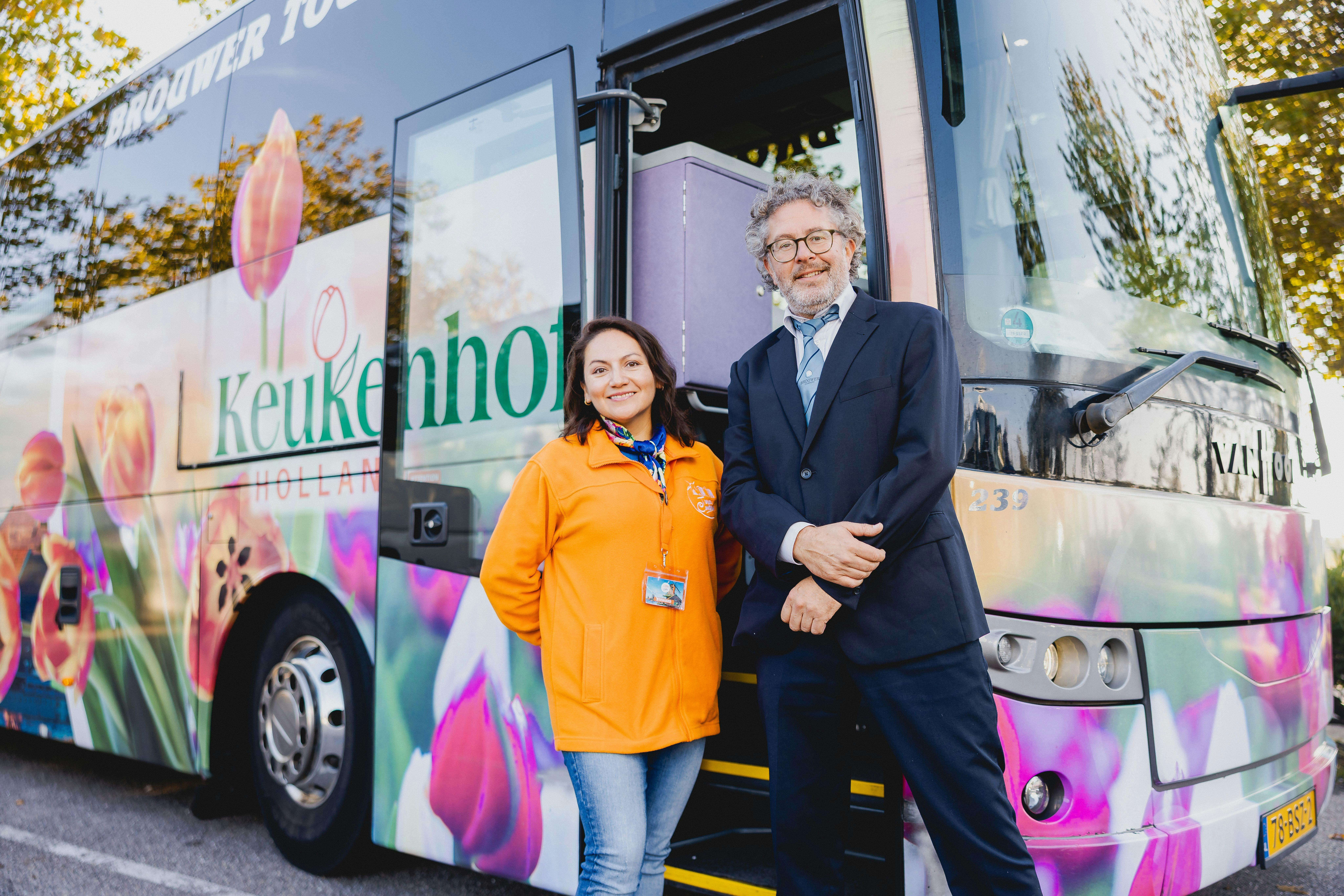 Δύο άτομα στέκονται μπροστά από ένα πολύχρωμο λεωφορείο διακοσμημένο με εικόνες τουλίπας. Ο ένας φοράει πορτοκαλί σακάκι και ο άλλος μαύρο κοστούμι.