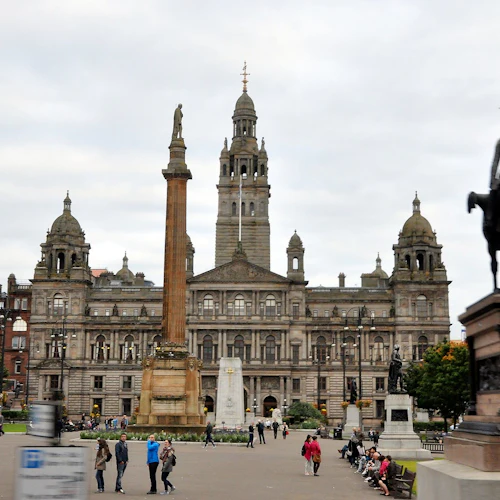 Människor som går och sitter på ett torg med statyer och en historisk byggnad i bakgrunden. En staty bär en trafikkon.