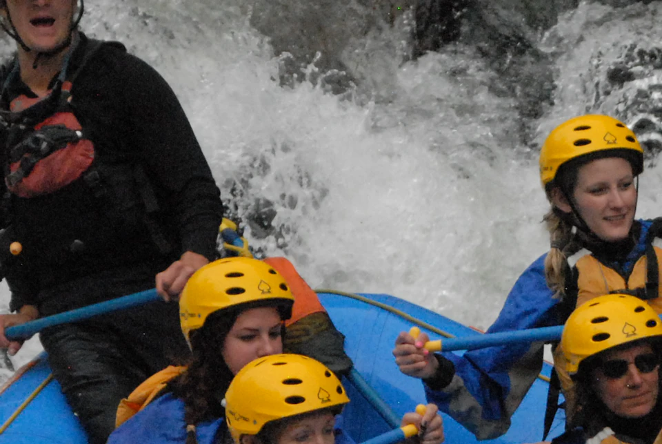 A group of people in yellow helmets are white-water rafting in a blue inflatable raft, navigating through rocky rapids.