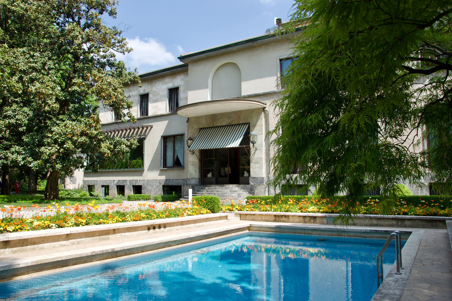 A two-story house with striped awnings, surrounded by a garden with bright flowers, and a swimming pool in the foreground.