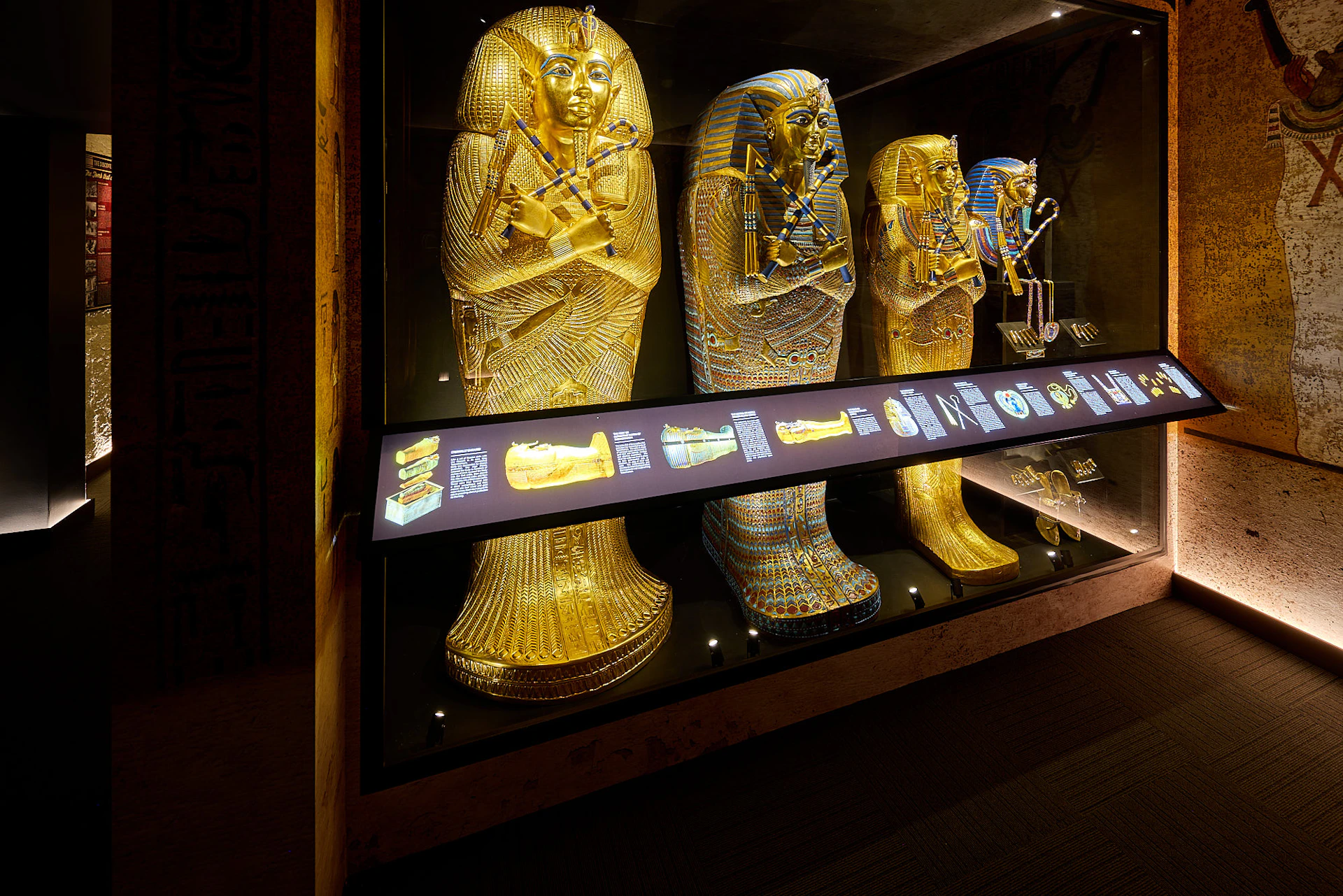 Three illuminated Egyptian sarcophagi displayed in a museum exhibit, with an informational panel in front of them.