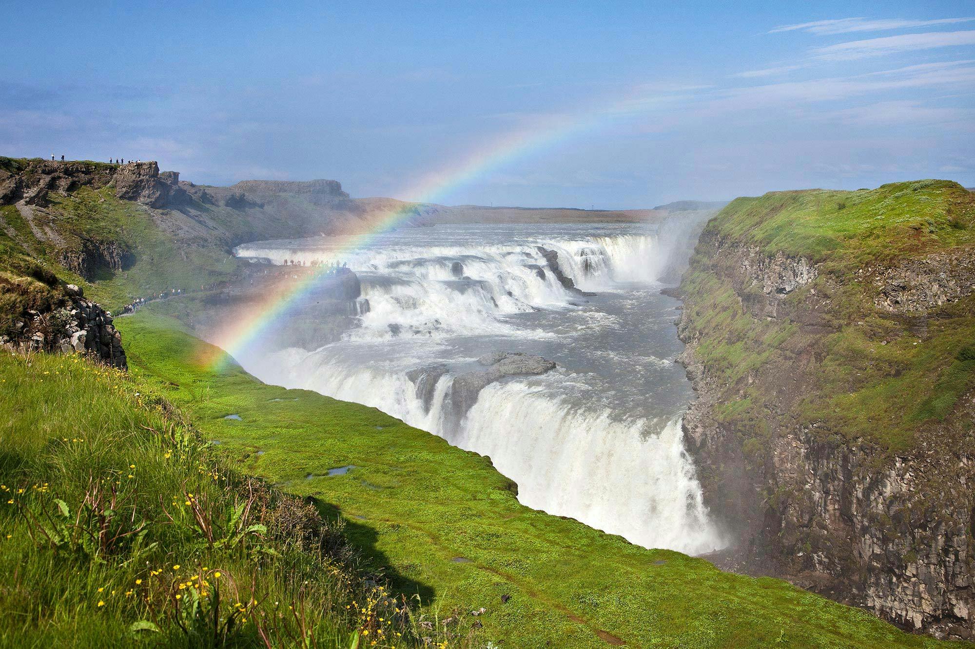 A waterfall cascades down tiers surrounded by green cliffs, with a vivid rainbow arching across the misty scene under a blue sky.