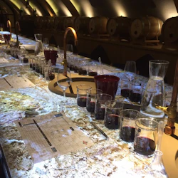 En bordsduk för en vinprovning med bägare av rött vin, glas, karaffer och former i ett svagt upplyst rum med träfat i bakgrunden.