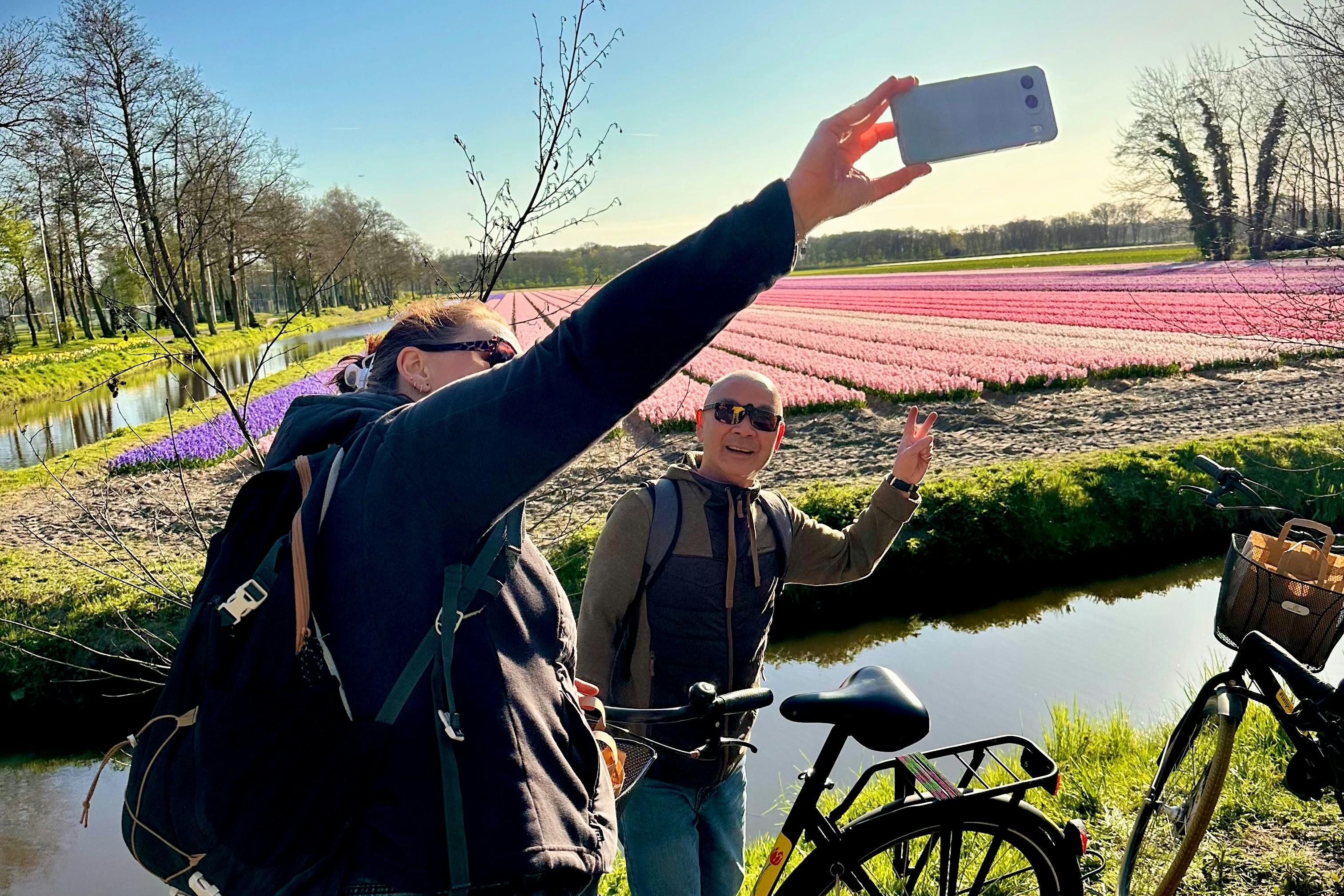 Two people with bicycles take a selfie in front of a field of blooming pink flowers, a clear sky, and a small canal.