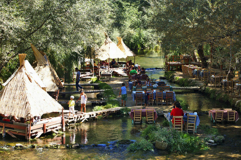 Outdoor dining area with wooden tables and chairs set in a shallow stream, surrounded by trees and thatched huts. People are seated and walking around.