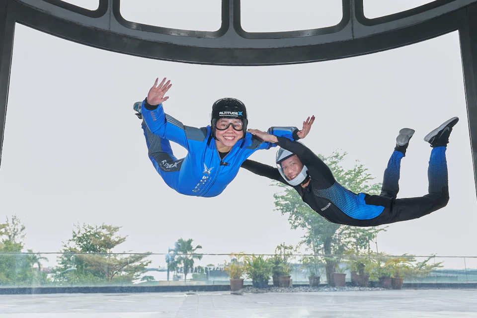 Two people in flight suits are flying inside a vertical wind tunnel with transparent walls, surrounded by branding logos.