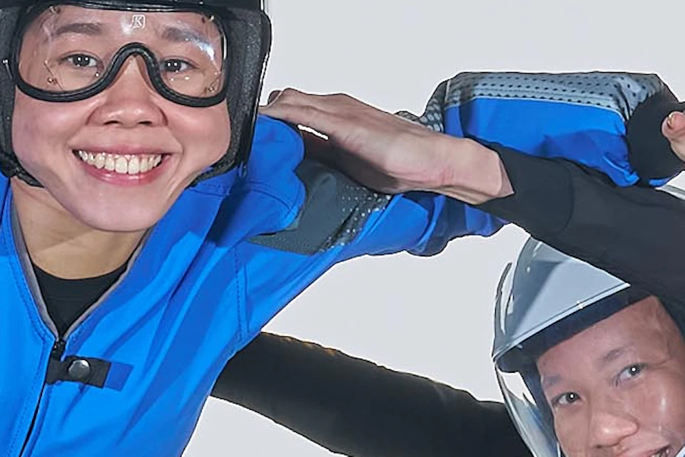 Two people in flight suits are flying inside a vertical wind tunnel with transparent walls, surrounded by branding logos.