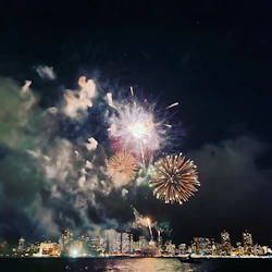 Fireworks exploding in the night sky above a city skyline with reflections on the water.
