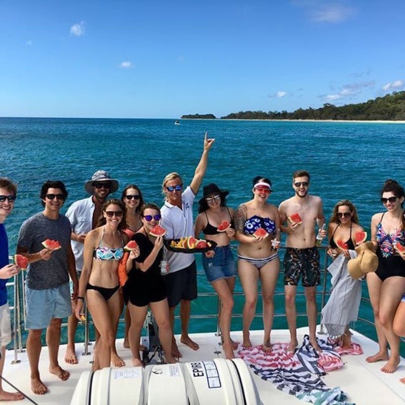 Μια ομάδα ανθρώπων με μαγιό που κρατούν φέτες καρπουζιού σε μια βάρκα μια ηλιόλουστη μέρα, με φόντο τη θάλασσα και τις μακρινές ακτές.