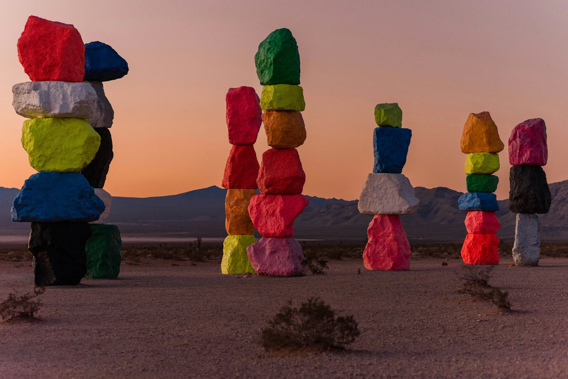 Brightly colored stacked rock formations in a desert landscape during sunset, with mountains in the background.