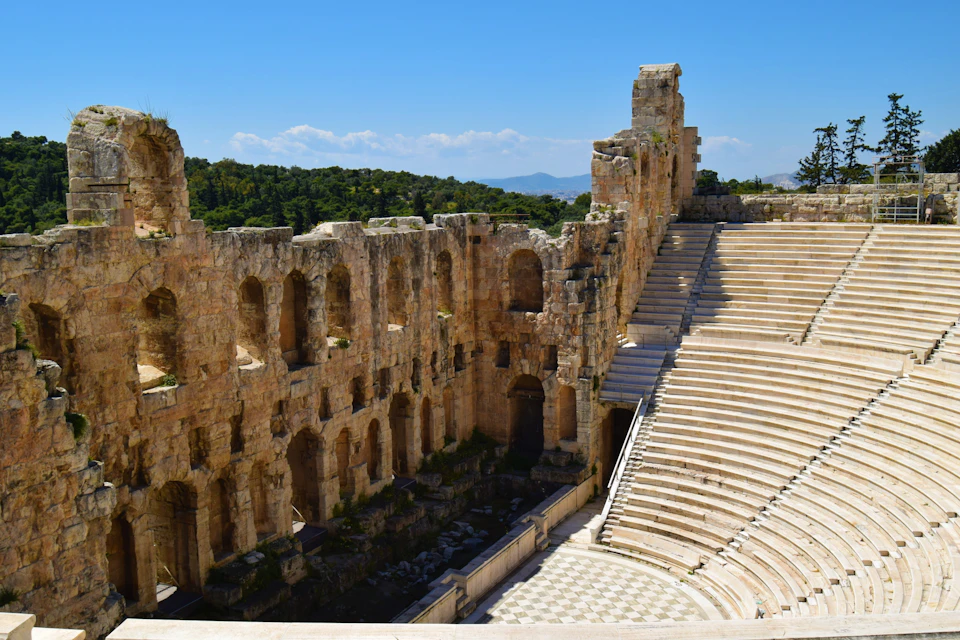 An ancient stone amphitheater with tiered seating and arched walls under a clear blue sky, surrounded by greenery.