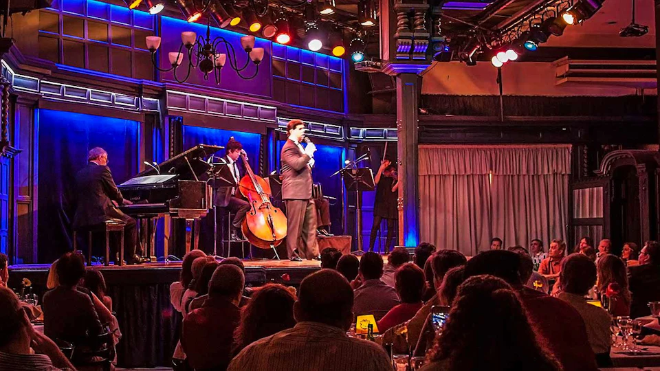 Man speaking on stage with a jazz band, including a double bass, piano, and saxophone, before an audience in dim lighting.