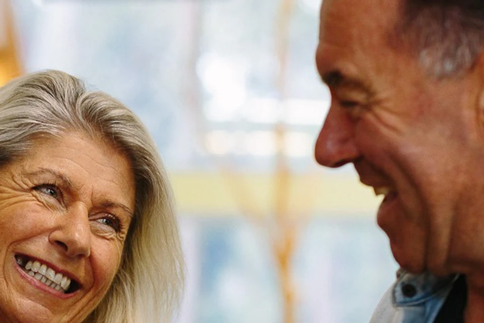 Two people smiling at each other indoors; one holds a stuffed koala. A rack of items and signage are visible in the background.
