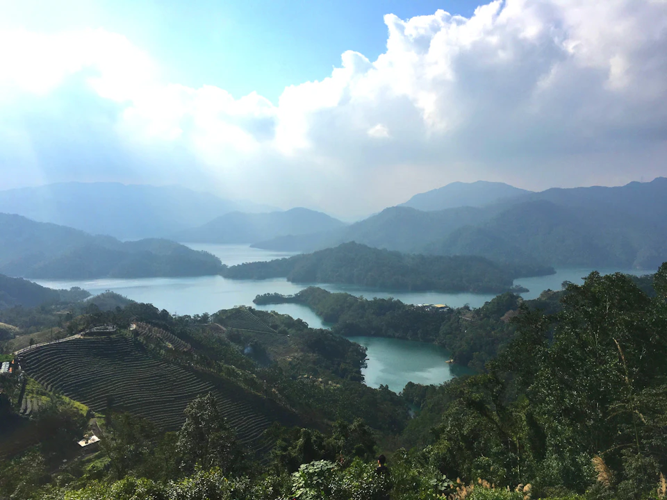 A serene landscape with tree-covered hills, a lake with islands, and distant mountains under a partly cloudy sky.
