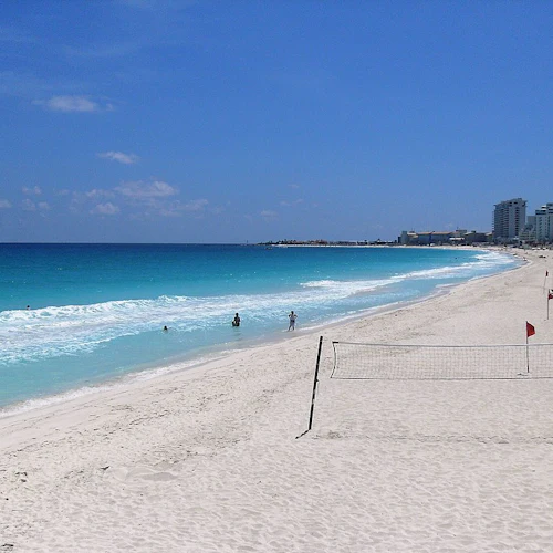 Sandstrand mit schwimmenden und spazierenden Menschen, Meereswellen, Volleyballnetz, Jetskis und hohen Gebäuden im Hintergrund unter blauem Himmel.