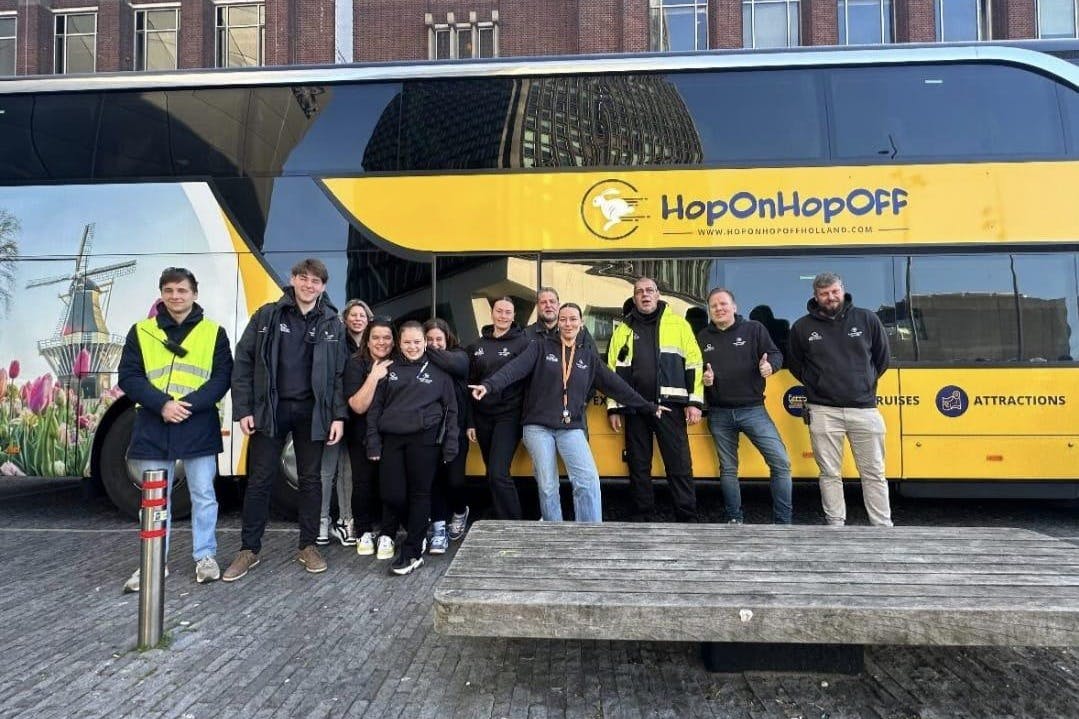 A group of people posing in front of a tour bus with a windmill and tulip mural, some wearing high-visibility jackets.