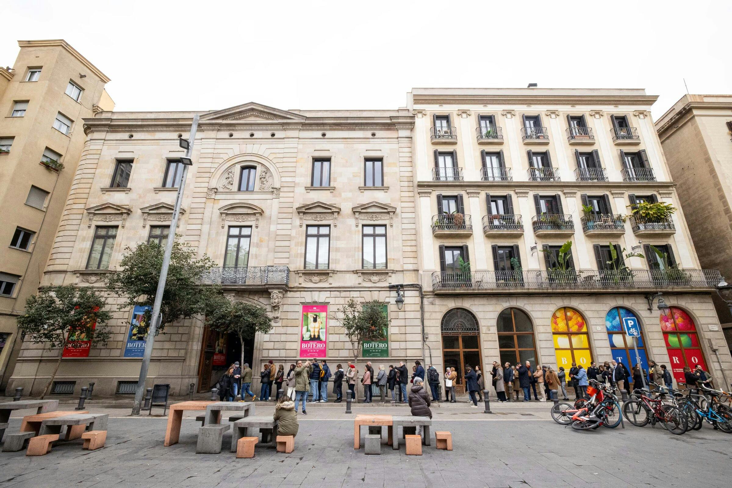 A group of people stands in line outside a classical-style building with large windows and colorful posters on its facade.