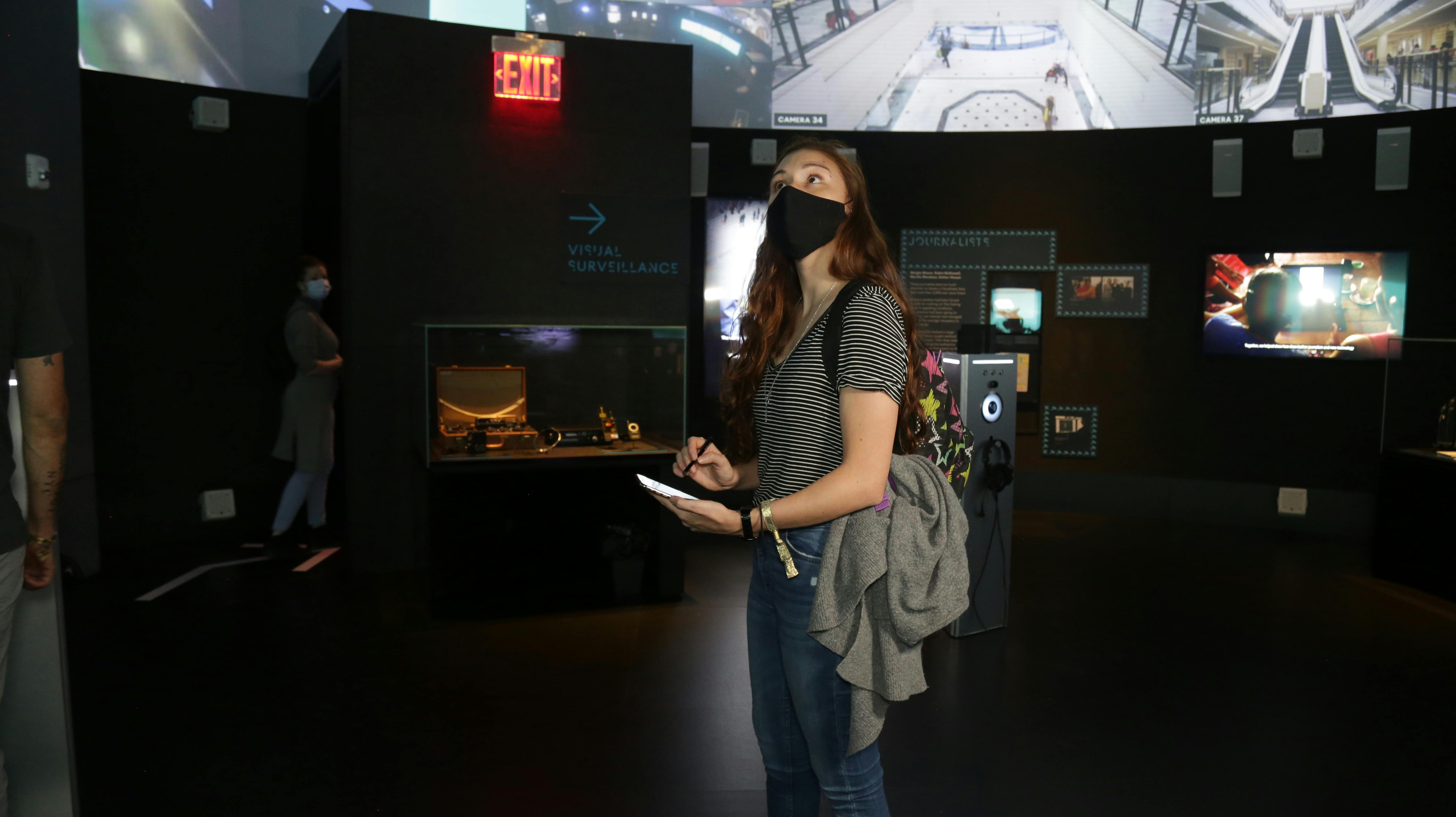 Μια γυναίκα με ριγέ πουκάμισο και μαύρη μάσκα κρατά σημειώσεις σε έναν εκθεσιακό χώρο του μουσείου, κοντά σε μια προθήκη και μια πινακίδα «ΕΞΟΔΟΥ».