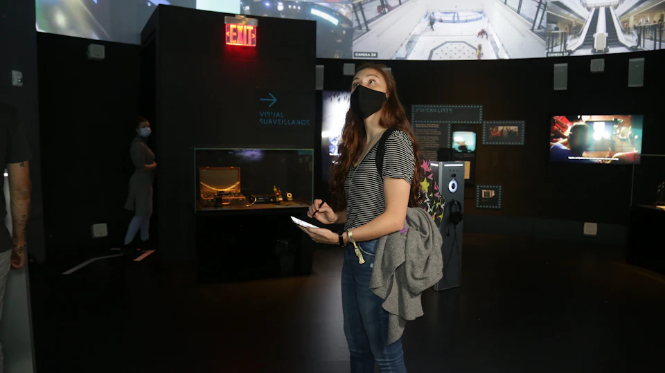 A woman in a striped shirt and black mask takes notes in a museum exhibit area, near a display case and an "EXIT" sign.