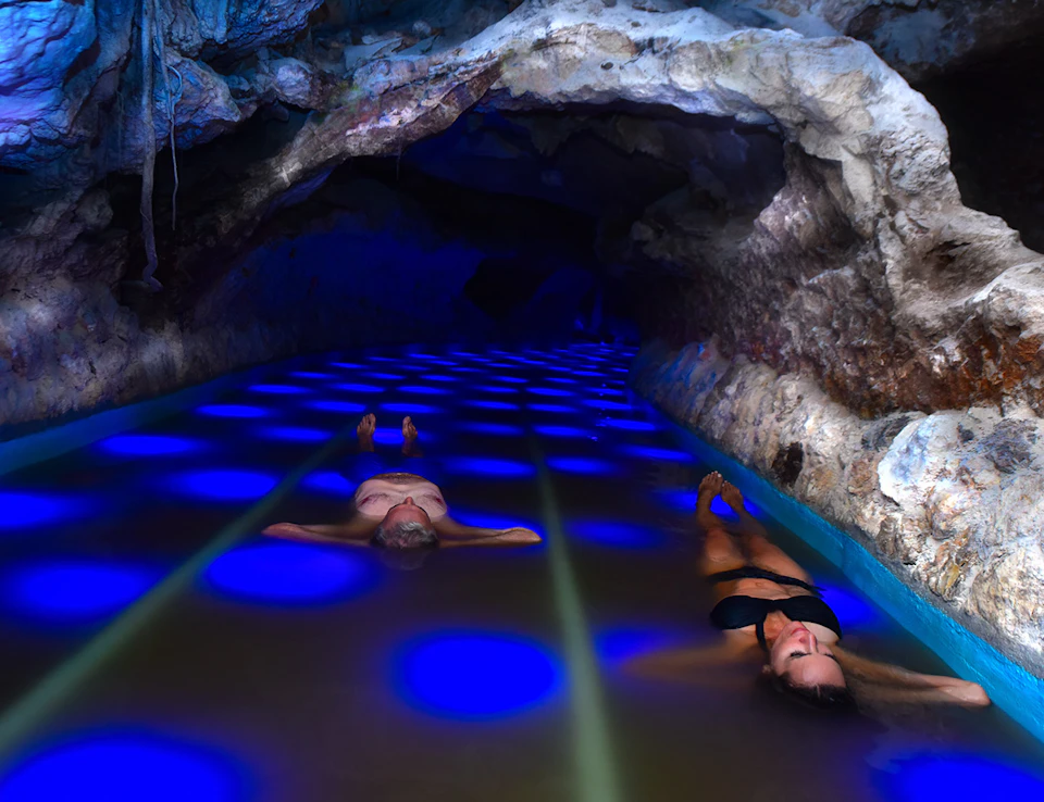 Two people float on their backs in a cave pool illuminated with blue lights, surrounded by rocky walls.