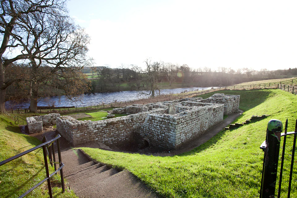 Ancient stone ruins near a river, surrounded by grass and trees, viewed from descending stone steps.