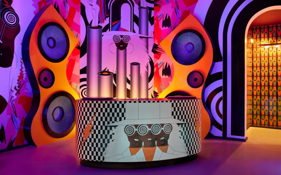A colorful art installation featuring cylindrical structures, abstract patterns, and stylized human faces on a checkered counter.