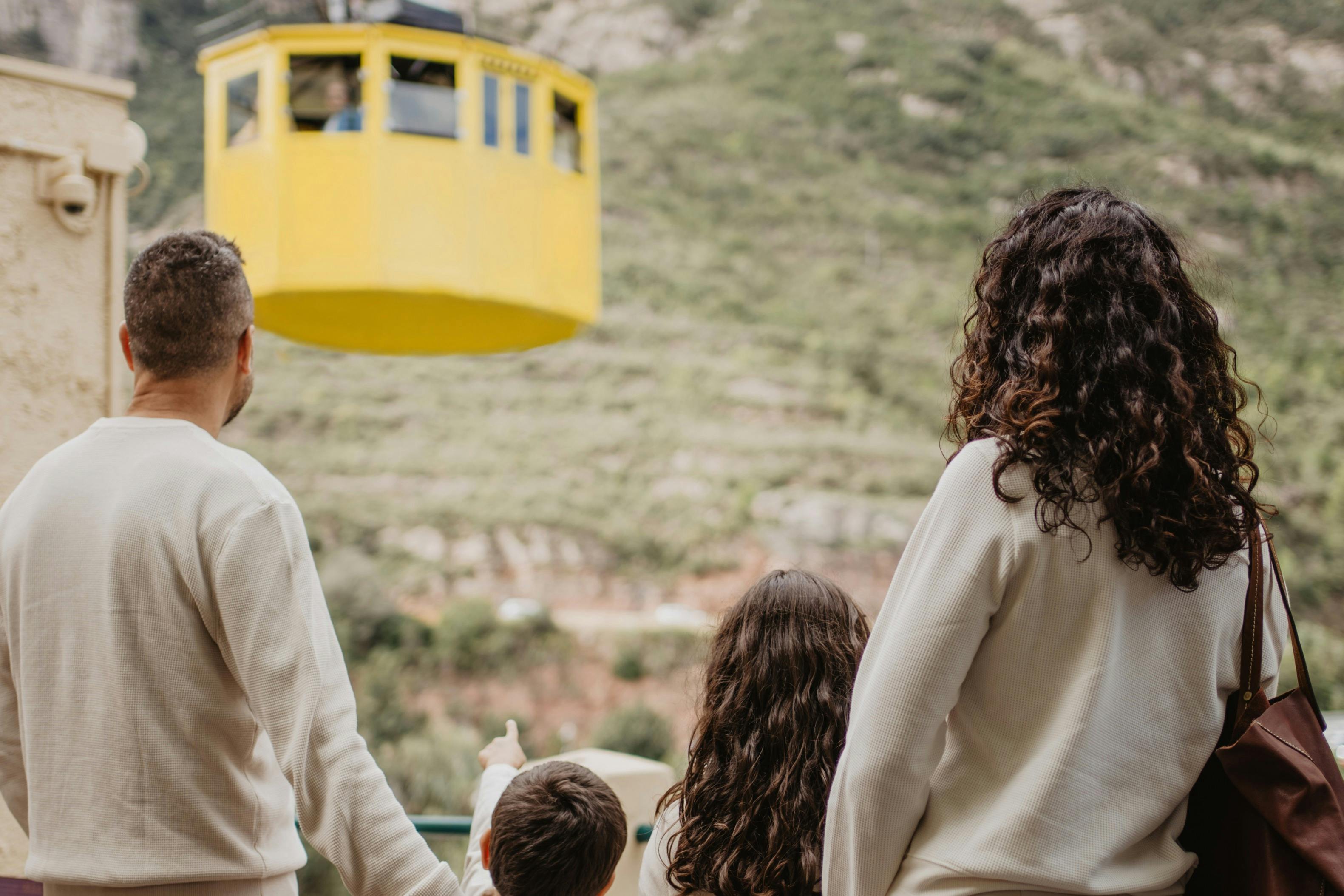 Una familia de cuatro miembros observa un teleférico amarillo que asciende por la ladera verde de una montaña.