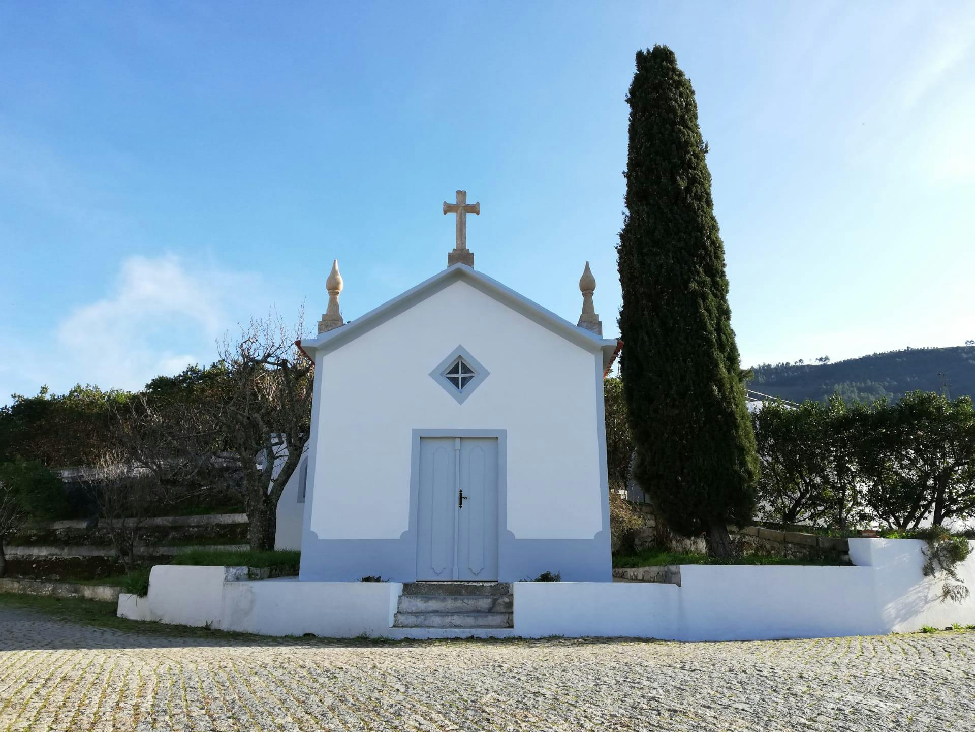 Een kleine witte kapel met een kruis erop, een blauwe deur en een hoge cipres ernaast.