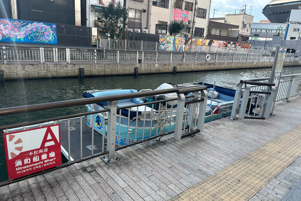A small docked boat by a canal, with a metal railing and a brick walkway in the foreground, and buildings in the background.