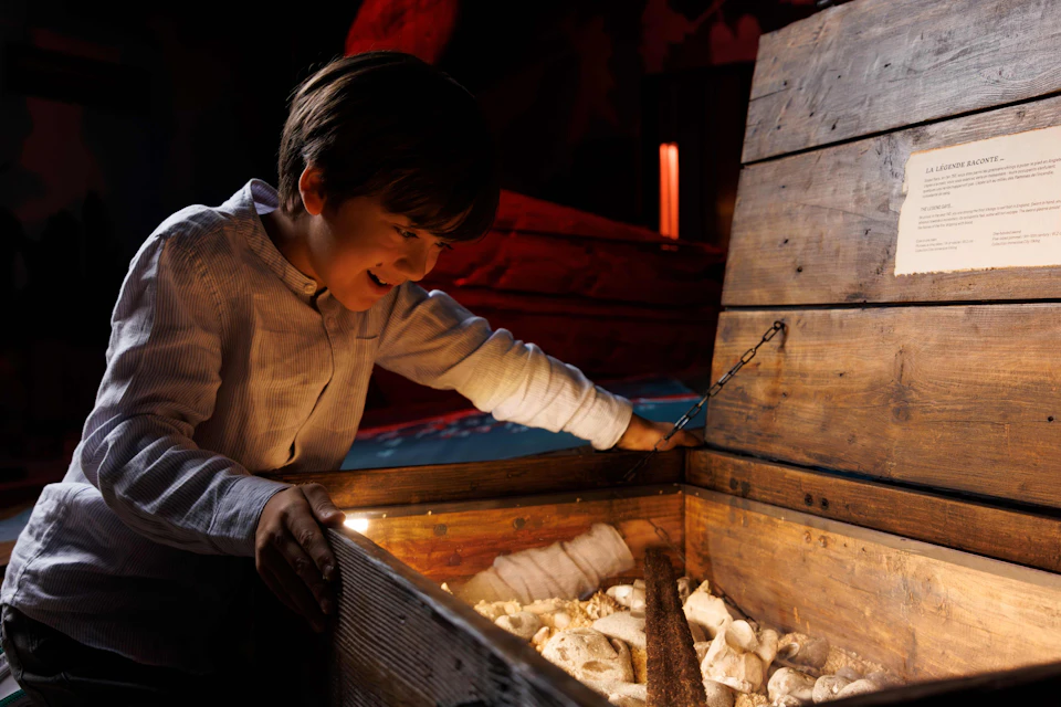 A young boy excitedly looks into an open wooden chest filled with various objects, illuminated warmly in a dimly lit setting.