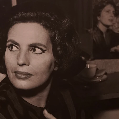 En kvinna med dramatisk ögonmakeup och kort mörkt hår tittar åt sidan; en kopp och ett fat sitter på bordet bakom henne.